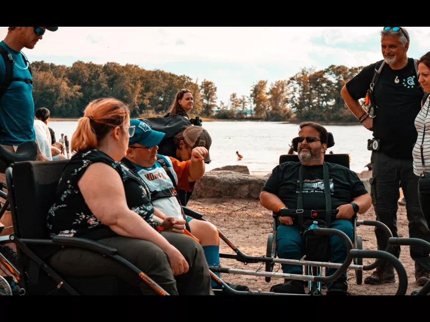 Au bord d'un lac tranquille, trois individus atteints de spina bifida se retrouvent en chaises roulantes pour échanger des moments de complicité. La quiétude du lieu contraste avec la résilience et la solidarité qui se dégagent de leur discussion.